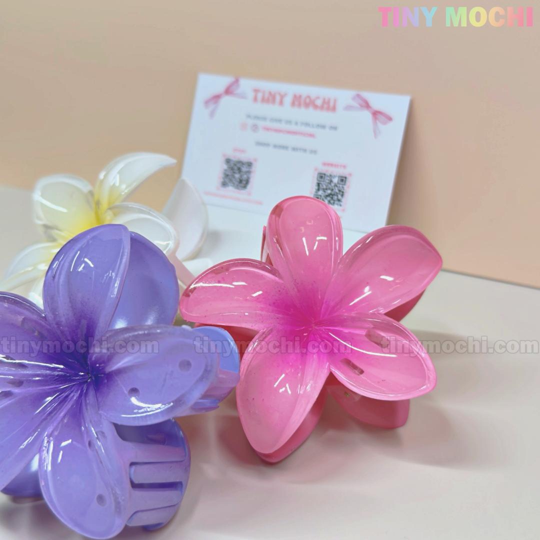 Three large Plumeria Flower Hair Claws by Tiny Mochi—colorful floral hair clips in purple, pink, and white—are displayed in front. A Tiny Mochi business card with QR codes appears blurred in the background. Stylish & strong grip for any hairstyle.