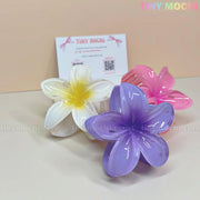 Three Tiny Mochi Plumeria Flower Hair Claws for women & girls in white, purple, and pink are shown on a light surface with a business card and Tiny Mochi watermark, highlighting these stylish summer hair accessories.