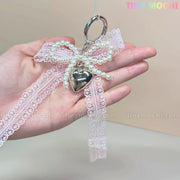 A hand holds a Kawaii Lace Bow Keychain with pearl beads and a silver heart charm by Tiny Mochi, perfect as a cute bag charm, phone strap, or car mirror accessory. Tiny Mochi appears in pastel colors in the upper right corner.