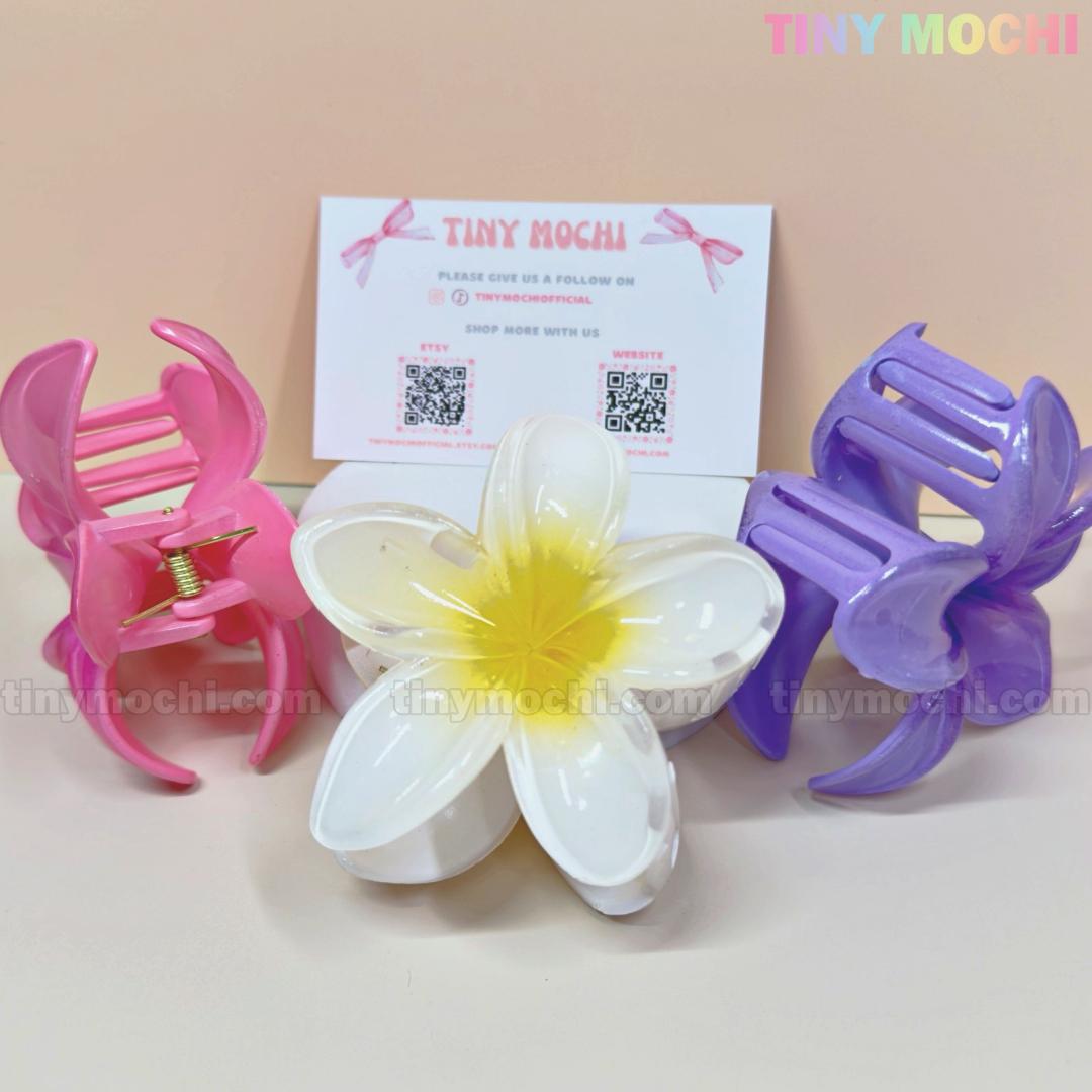 Three Plumeria Flower Hair Claws from Tiny Mochi—pink, white floral, and purple—are displayed in front of a Tiny Mochi card. These stylish summer hair clips for women stand out against the soft peach background.