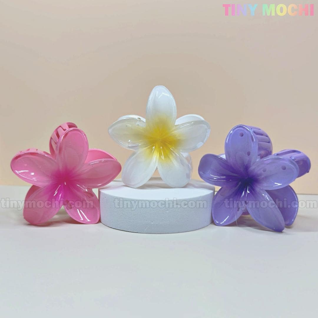 The Tiny Mochi Plumeria Flower Hair Claws—pink, white with yellow center, and purple—are displayed on a white pedestal against a pastel backdrop. These colorful floral hair clips offer stylish summer flair and strong hold for women and girls.