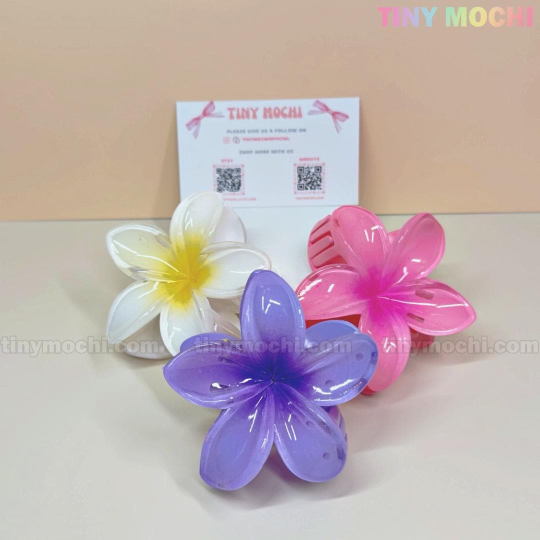 Three Plumeria Flower Hair Claws by Tiny Mochi, in white, pink, and purple, are shown in front of a TINY MOCHI card on a light beige surface. These colorful floral clips offer a strong grip for stylish summer hairdos.