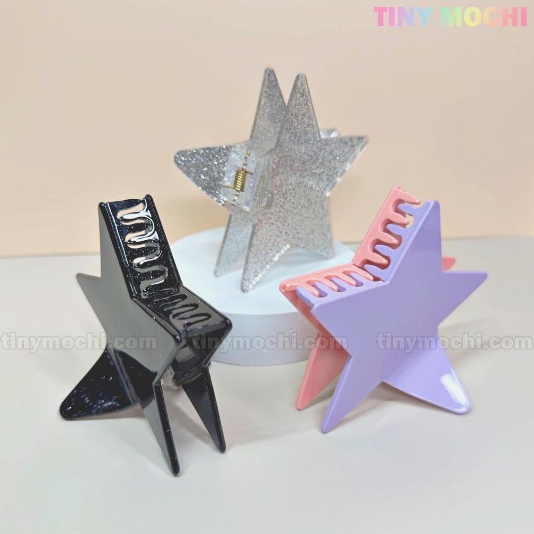 Three large Glitter Star Hair Claw Clips by Tiny Mochi—black, silver glitter, and pastel pink-purple—stand on a white platform. The pastel pink background with the TINY MOCHI logo highlights these Y2K aesthetic acetate hair accessories.