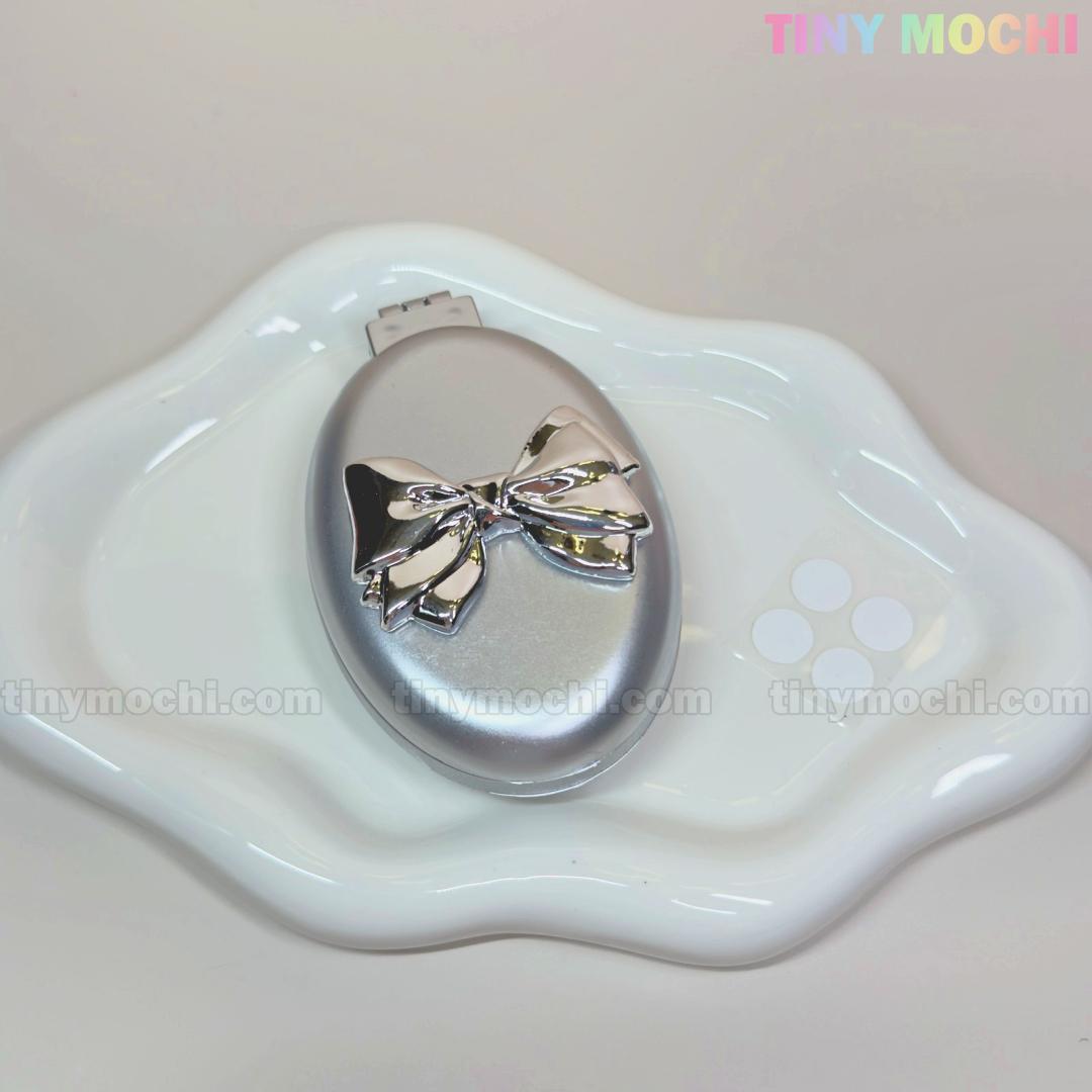 The Oval Silver Folding Travel Mirror Comb by Tiny Mochi, a compact pocket-sized hair brush with a built-in mirror, sits on a wavy-edged white dish. The pastel Tiny Mochi logo appears in the image corner.