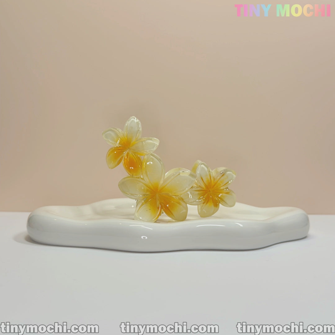 Three glossy Plumeria Flower Claw Clip Hair Accessories by Tiny Mochi, in yellow and white, rest on a smooth wavy-edged ceramic tray against a neutral background. “TINYMochi.com” appears at the bottom and top right corner.