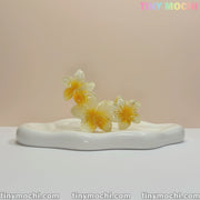 Three glossy Plumeria Flower Claw Clip Hair Accessories by Tiny Mochi, in yellow and white, rest on a smooth wavy-edged ceramic tray against a neutral background. “TINYMochi.com” appears at the bottom and top right corner.