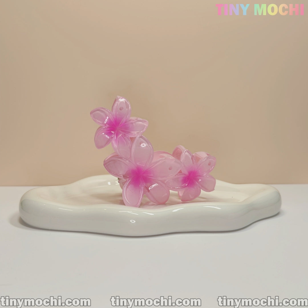 Three translucent pink plumeria flower claw clips by Tiny Mochi rest on a glossy white dish. These elegant, non-slip hair accessories are perfect for women and kids, adding a delicate floral touch to any hairstyle.