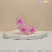 Three translucent pink plumeria flower claw clips by Tiny Mochi rest on a glossy white dish. These elegant, non-slip hair accessories are perfect for women and kids, adding a delicate floral touch to any hairstyle.