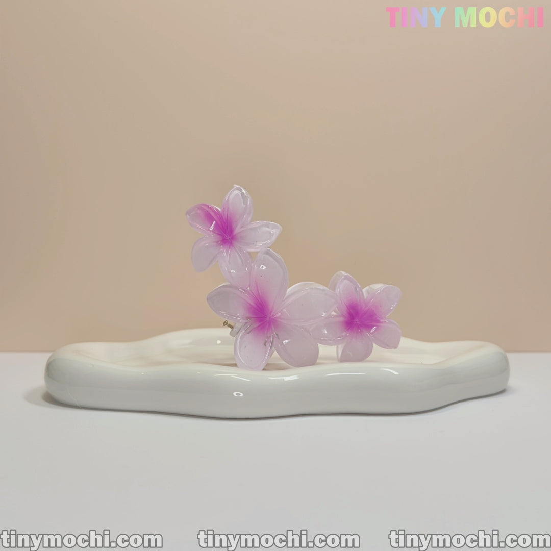A translucent pink and purple Plumeria Flower Claw Clip Hair Accessory by Tiny Mochi sits on a wavy white ceramic tray, with the brand name in pastel at the top right. Perfect for women & kids, this durable non-slip clip suits thick or thin hair.