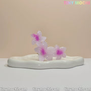 A translucent pink and purple Plumeria Flower Claw Clip Hair Accessory by Tiny Mochi sits on a wavy white ceramic tray, with the brand name in pastel at the top right. Perfect for women & kids, this durable non-slip clip suits thick or thin hair.