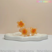 Three orange glass sculptures, shaped like the Plumeria Flower Claw Clip Hair Accessory by Tiny Mochi, are arranged on a white wavy ceramic base. TINY MOCHI appears in pastel colors at the top-right against a plain background.