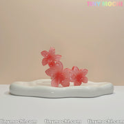 Three translucent pink flower-shaped candies are arranged on a wavy-edged dish, evoking the Plumeria Flower Claw Clip Hair Accessory by Tiny Mochi. “TINY MOCHI” appears in pastel at the top right corner.