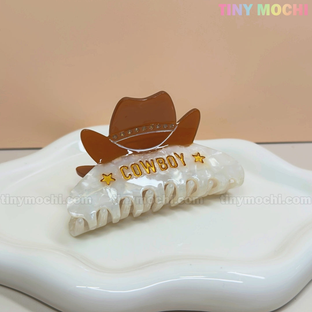 The Tiny Mochi Western Cowboy Hair Claw Clip features a white hat with COWBOY and yellow stars, perfect for a cowgirl look. Shown on a ceramic tray with a beige background and pastel Tiny Mochi logo in the top right corner.