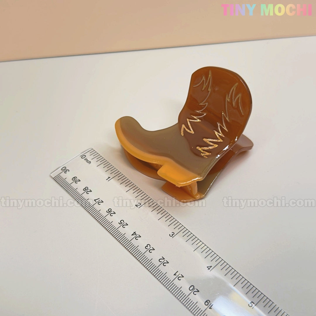 The Tiny Mochi Western Cowboy Hair Claw Clip—shaped like a brown boot with gold accents—rests by a ruler on a white background. Tiny Mochi appears in pastel at the top right, and the clip is about 5 cm long.