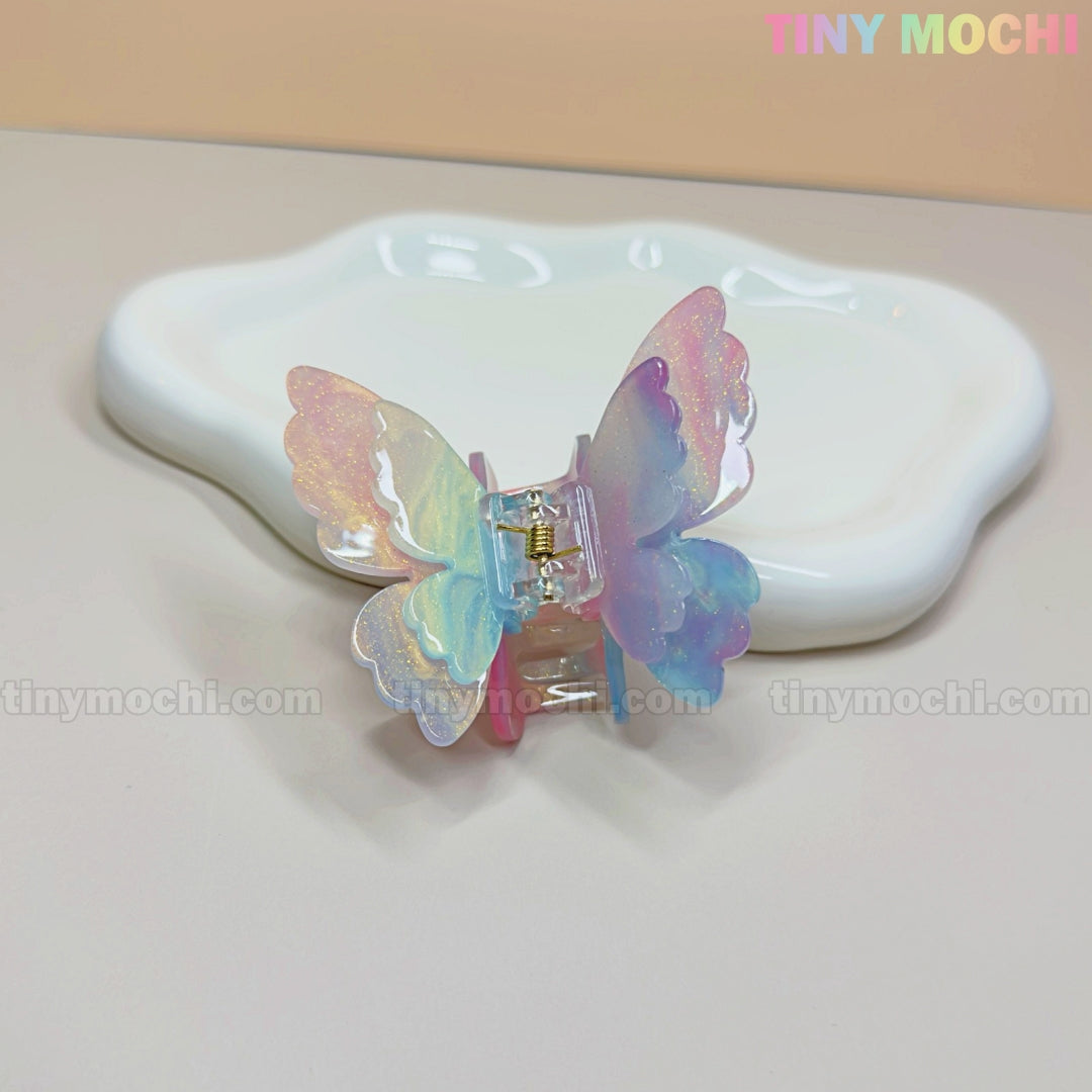 The NEW UV Color-Changing Butterfly Hair Claw Clip by Tiny Mochi shines with pastel, double-layer acetate wings in front of a white dish on beige—colorful Tiny Mochi text at top right, perfect for a magical Spring Fairycore vibe.