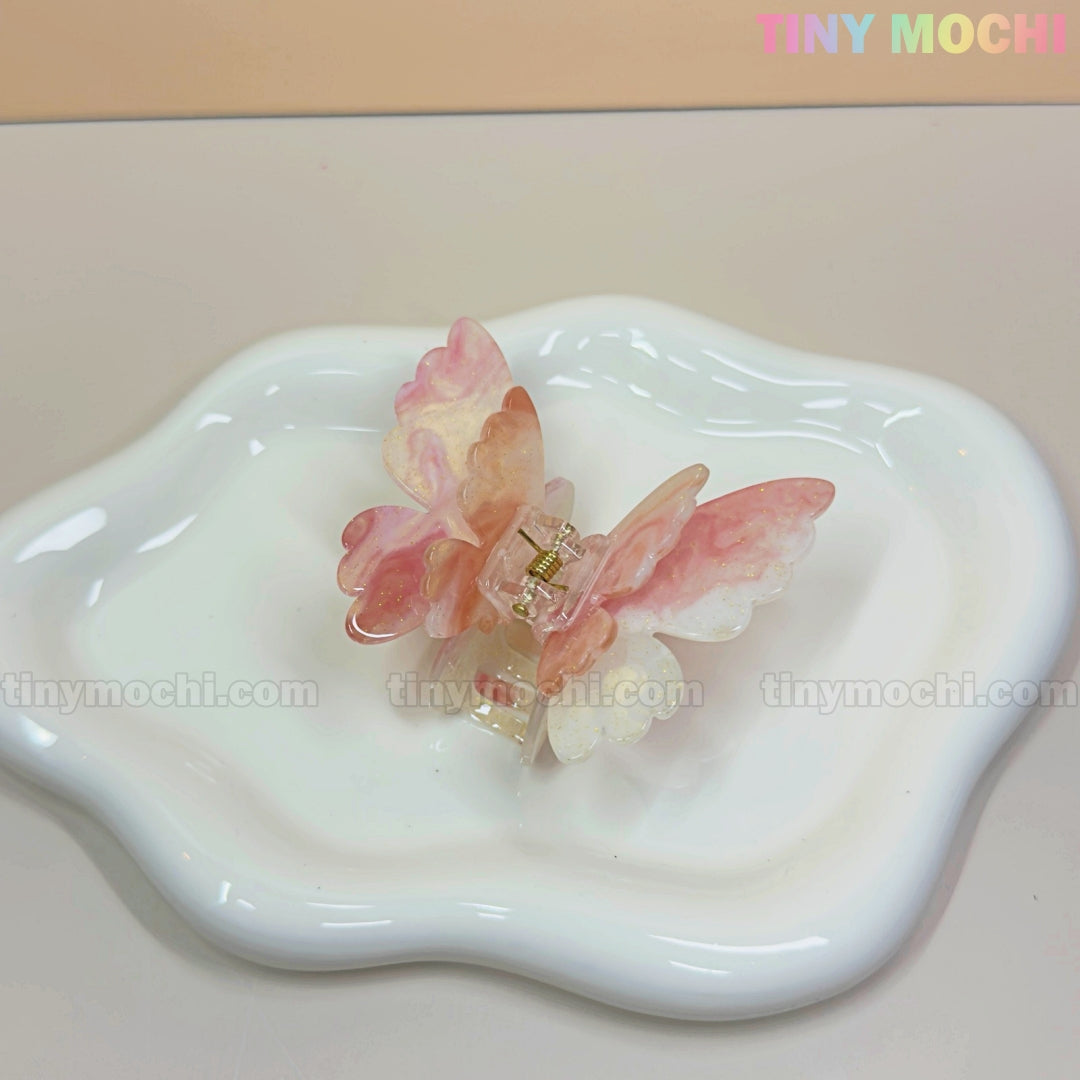 A NEW UV Color-Changing Butterfly Hair Claw Clip—Double Layer Acetate Pastel Hair Accessory by Tiny Mochi rests on a wavy white ceramic dish. A pastel TINY MOCHI logo appears in the top right over a light beige background.