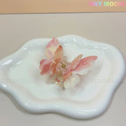 A NEW UV Color-Changing Butterfly Hair Claw Clip—Double Layer Acetate Pastel Hair Accessory by Tiny Mochi rests on a wavy white ceramic dish. A pastel TINY MOCHI logo appears in the top right over a light beige background.