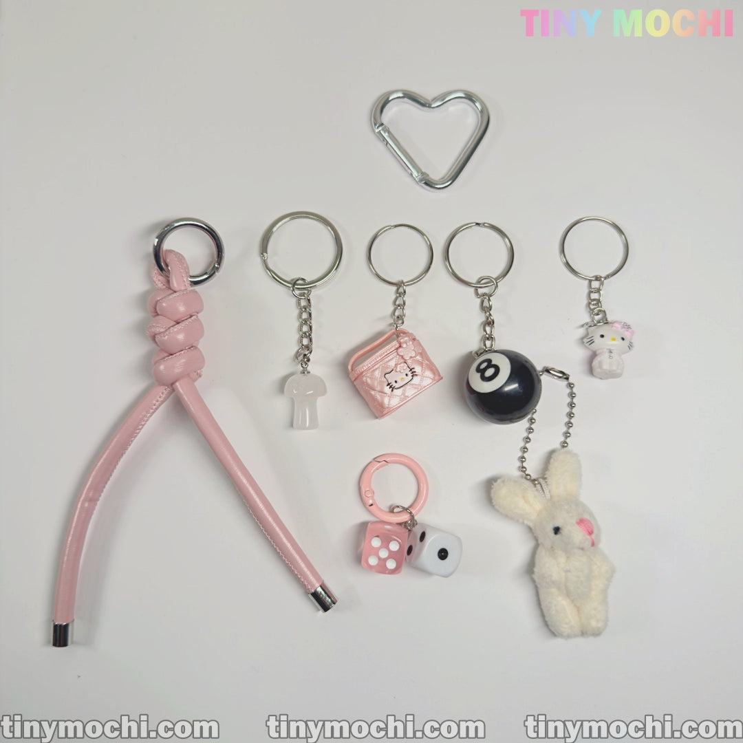The Tiny Mochi Cute Bunny and Charm Keychain features eight kawaii accessories, including a pink braided strap, heart-shaped ring, Y2K lock, 8-ball, dice, plush bunny, and two cute character charms—perfect for purses and backpacks.