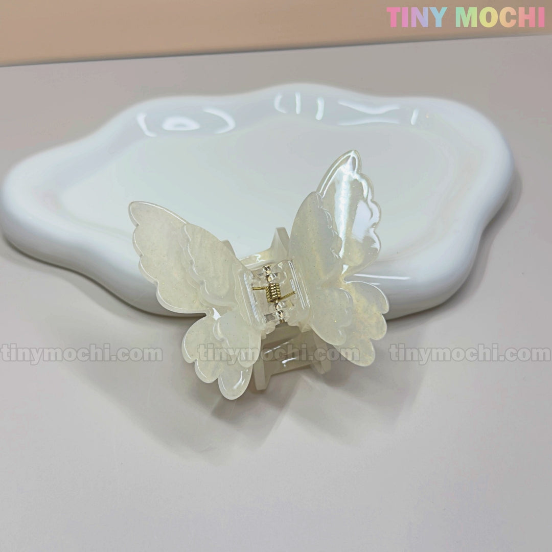The NEW UV Color-Changing Butterfly Hair Claw Clip—Double Layer Acetate Pastel Hair Accessory by Tiny Mochi sits on a wavy-edged white ceramic dish, with a light beige background and Tiny Mochi in pastel colors in the upper right corner.