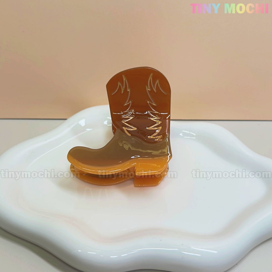 A glossy brown Western Cowboy Hair Claw Clip by Tiny Mochi, shaped like a boot, rests on a wavy-edged ceramic dish against a light beige background. “TINY MOCHI” in pastel colors adds a fun cowgirl vibe.