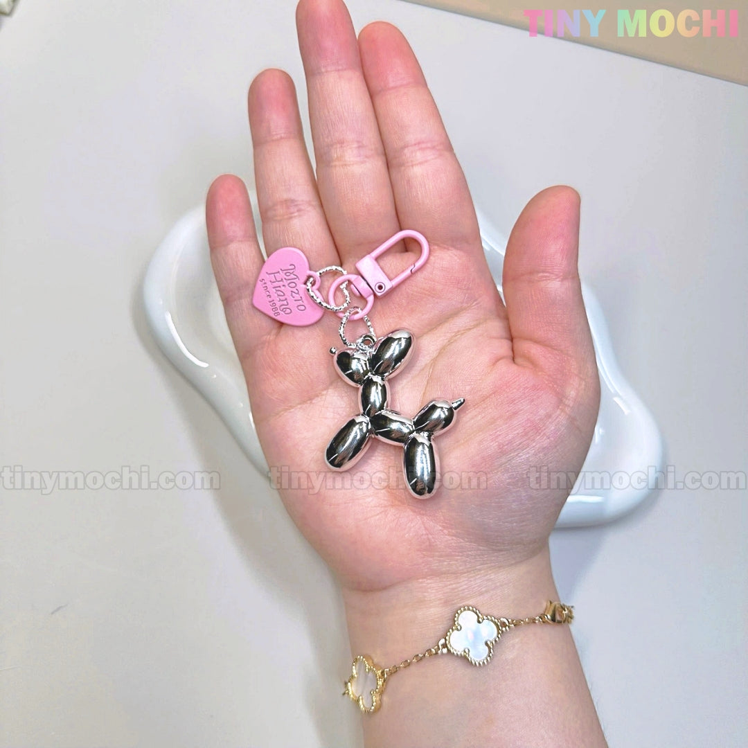 A hand holds the Tiny Mochi Balloon Dog Keychain Chrome Silver Bag Charm with a pink heart tag. The person wears a gold bracelet with white flower charms. Tiny Mochi appears in the corner.