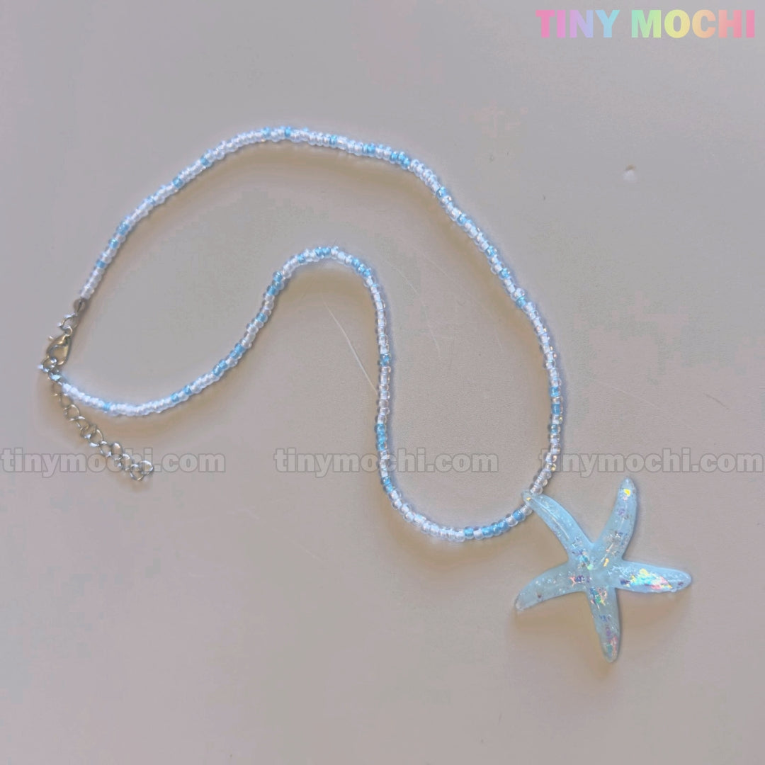 The Tiny Mochi Starfish Beach Necklace features a handmade iridescent blue pendant and small beads—ideal ocean aesthetic summer jewelry for girls and teens. The words tiny mochi appear faintly on the light surface.