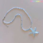 The Tiny Mochi Starfish Beach Necklace features a handmade iridescent blue pendant and small beads—ideal ocean aesthetic summer jewelry for girls and teens. The words tiny mochi appear faintly on the light surface.