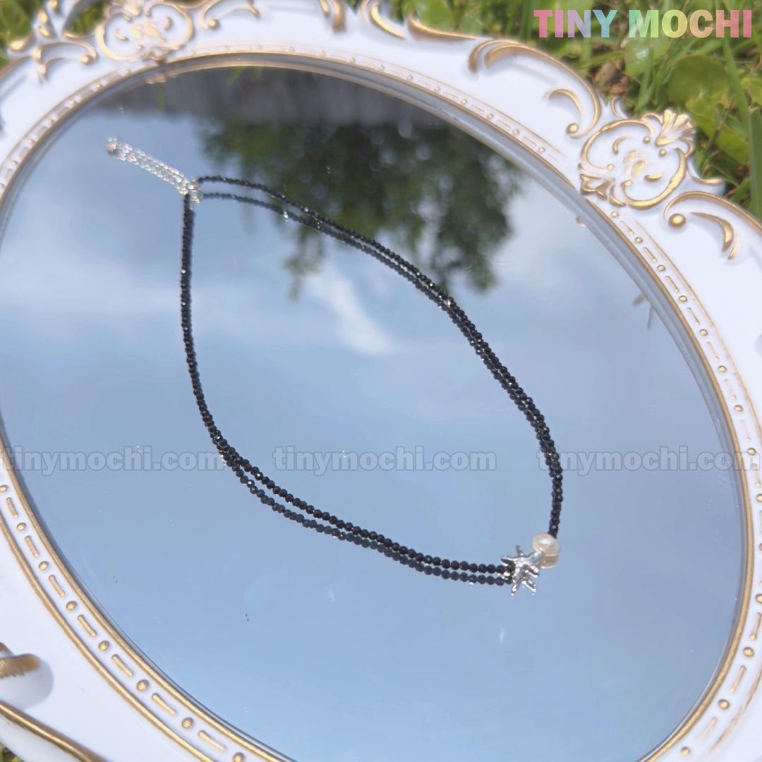 The Sterling Silver Starfish Necklace by Tiny Mochi features a handmade 925 silver charm and black beaded pearl choker, shown on an ornate mirror with reflections of greenery and sky. TINY MOCHI appears in the top right corner.