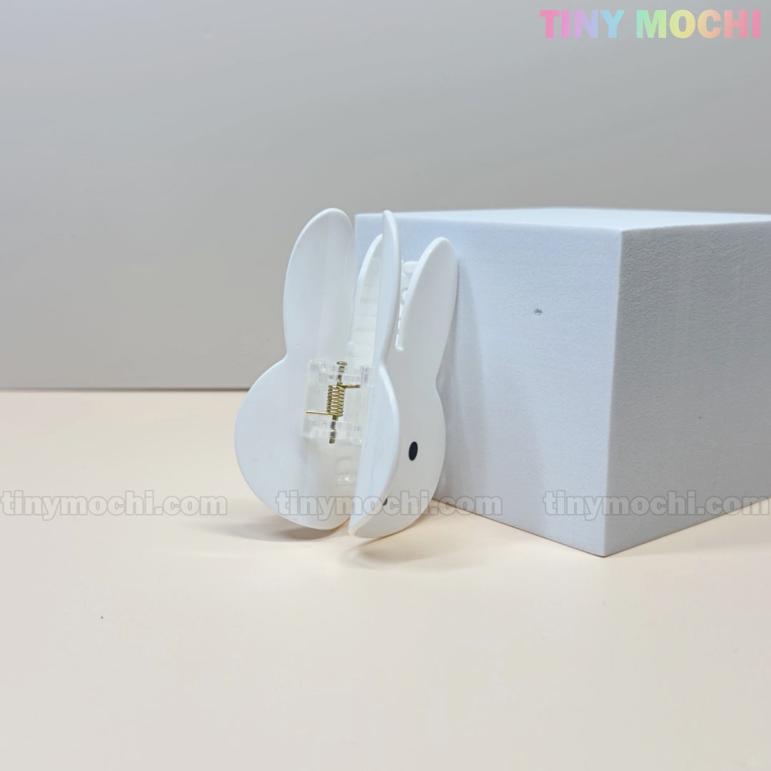 The Kawaii Bunny Face Hair Clip – White Rabbit Claw Clip by Tiny Mochi stands upright beside a white block on a neutral background, with TINY MOCHI in colorful letters at the top right and its metal spring visible.