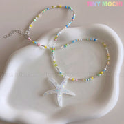 The Tiny Mochi Pastel Starfish Beach Bead Necklace features pastel blue, yellow, pink, and green beads with a handmade iridescent resin starfish pendant. Displayed on a white wavy-edge dish. Cute kawaii oceancore jewelry for girls and teens.