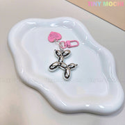 The Tiny Mochi Balloon Dog Keychain Chrome Silver Bag Charm features a pink heart tag and pink clip, adding Y2K-inspired style. It’s shown on a white ceramic dish with tinymochi.com faintly watermarked across the image.