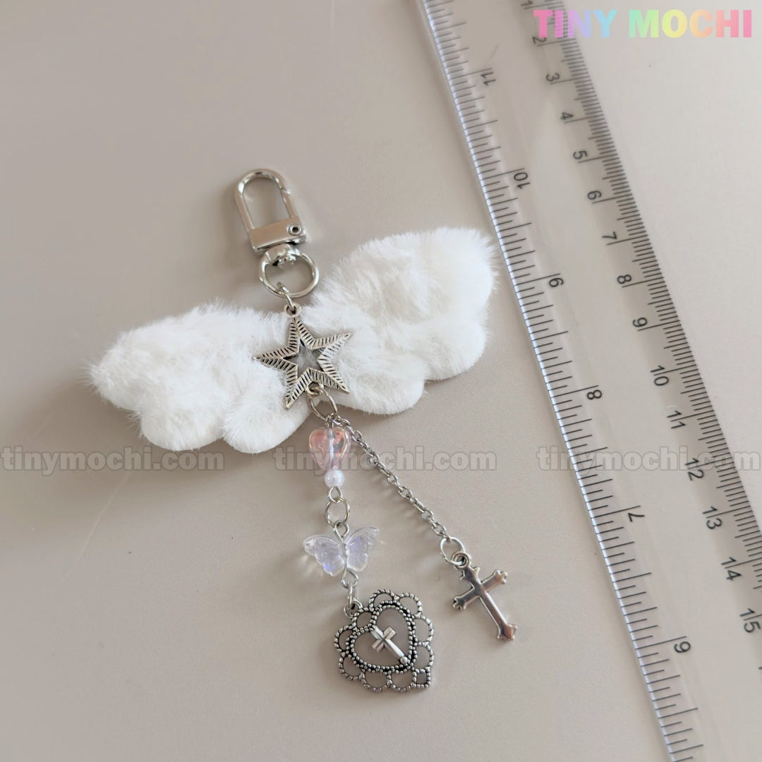 The Fluffy Angel Wing Keychain – Star & Cross Bag Charm Clip by Tiny Mochi features white wings and silver charms, channeling a soft gothic angel vibe. Shown near a clear ruler on a light surface, with “Tiny Mochi” in pastel at the top right.