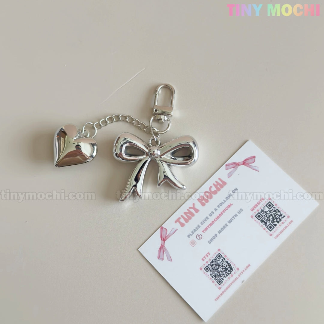 A Silver Bow Heart Keychain – Kawaii Mirror Bag Charm for Backpacks by Tiny Mochi rests beside a business card on a light surface, with the Tiny Mochi pastel logo shown in the top right corner.
