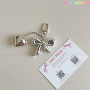 A Silver Bow Heart Keychain – Kawaii Mirror Bag Charm for Backpacks by Tiny Mochi rests beside a business card on a light surface, with the Tiny Mochi pastel logo shown in the top right corner.