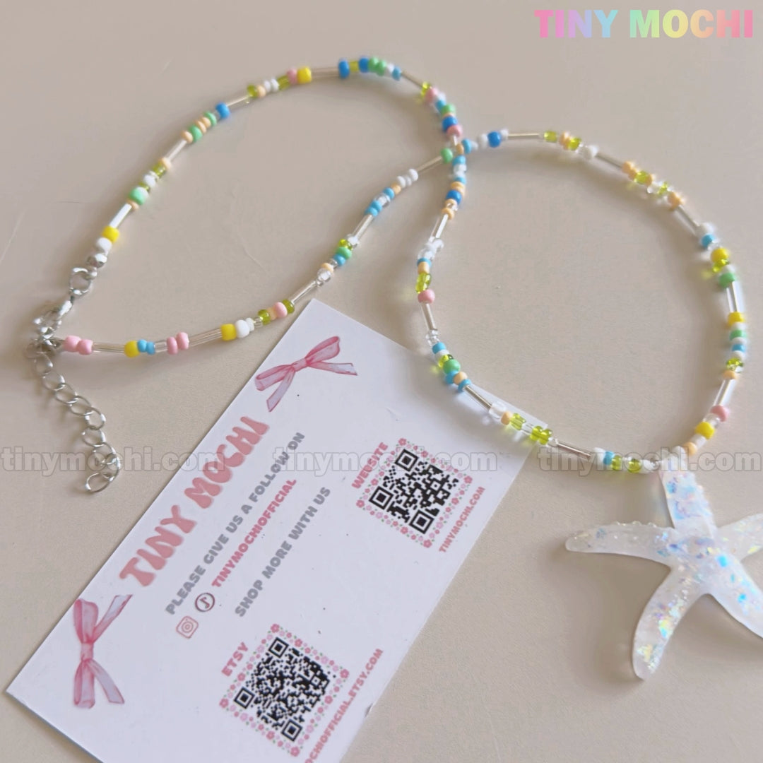 The Pastel Starfish Beach Bead Necklace by Tiny Mochi, a handmade iridescent resin choker for girls and teens, is shown beside a branded card with pink text, a bow illustration, and two QR codes on a light surface.