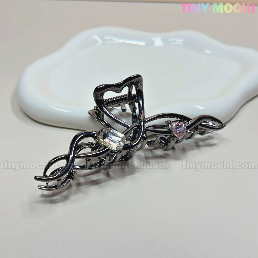 The Dark Silver Crystal Heart Hair Claw by Tiny Mochi—a black metal clip with a wavy design, clear and pink crystal heart accents—rests on a white ceramic dish. The background is neutral with the Tiny Mochi logo in the top right corner.