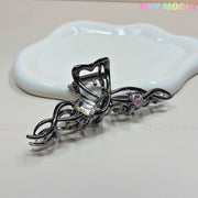 The Dark Silver Crystal Heart Hair Claw by Tiny Mochi—a black metal clip with a wavy design, clear and pink crystal heart accents—rests on a white ceramic dish. The background is neutral with the Tiny Mochi logo in the top right corner.