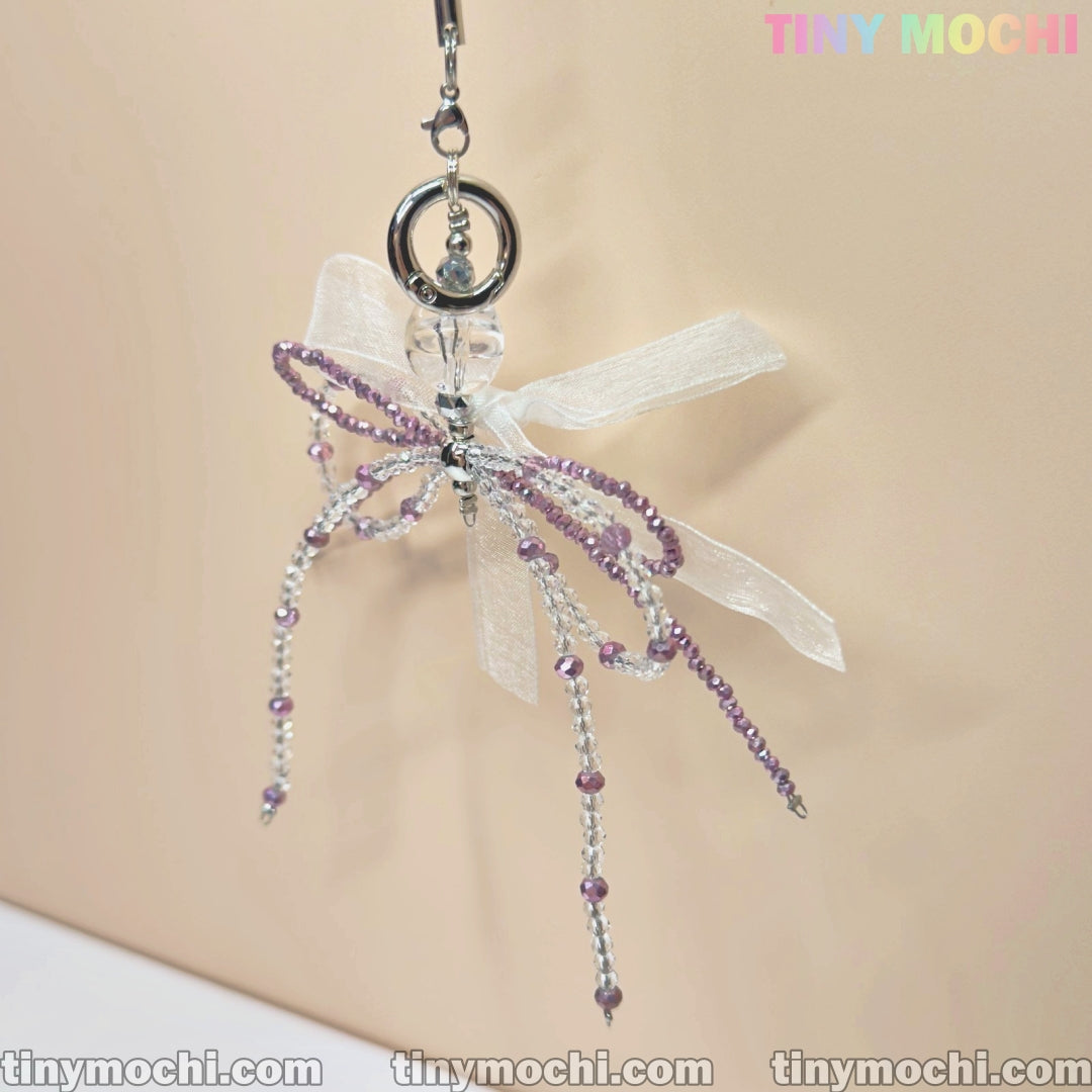 The Delicate Beaded Bow Charm – Elegant Y2K & Coquette Aesthetic Keychain from Tiny Mochi features a butterfly shape, purple and clear beads, and a sheer white ribbon bow, displayed against a light beige background.