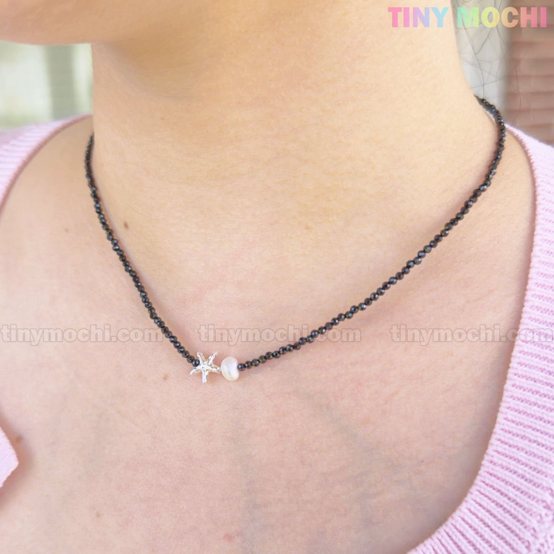 A person wearing the Tiny Mochi Sterling Silver Starfish Necklace—a handmade 925 silver charm on a black beaded pearl choker—styled with a light pink top.