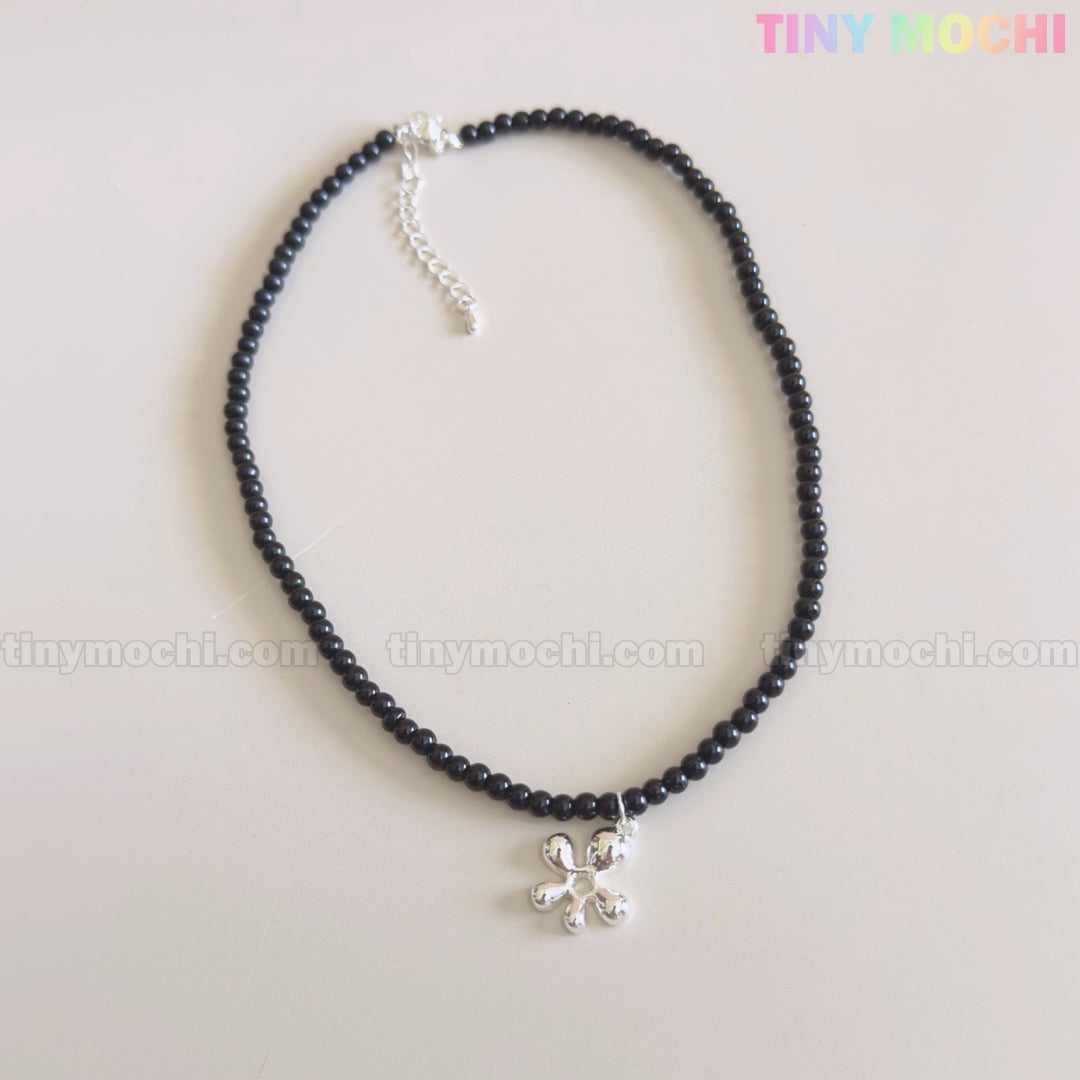 The Tiny Mochi Black Beaded Flower Necklace with Silver Floral Charm is a handmade Y2K choker, shown on a light background. The pastel Tiny Mochi logo appears in the top right corner. Perfect statement jewelry for her with soft grunge style.