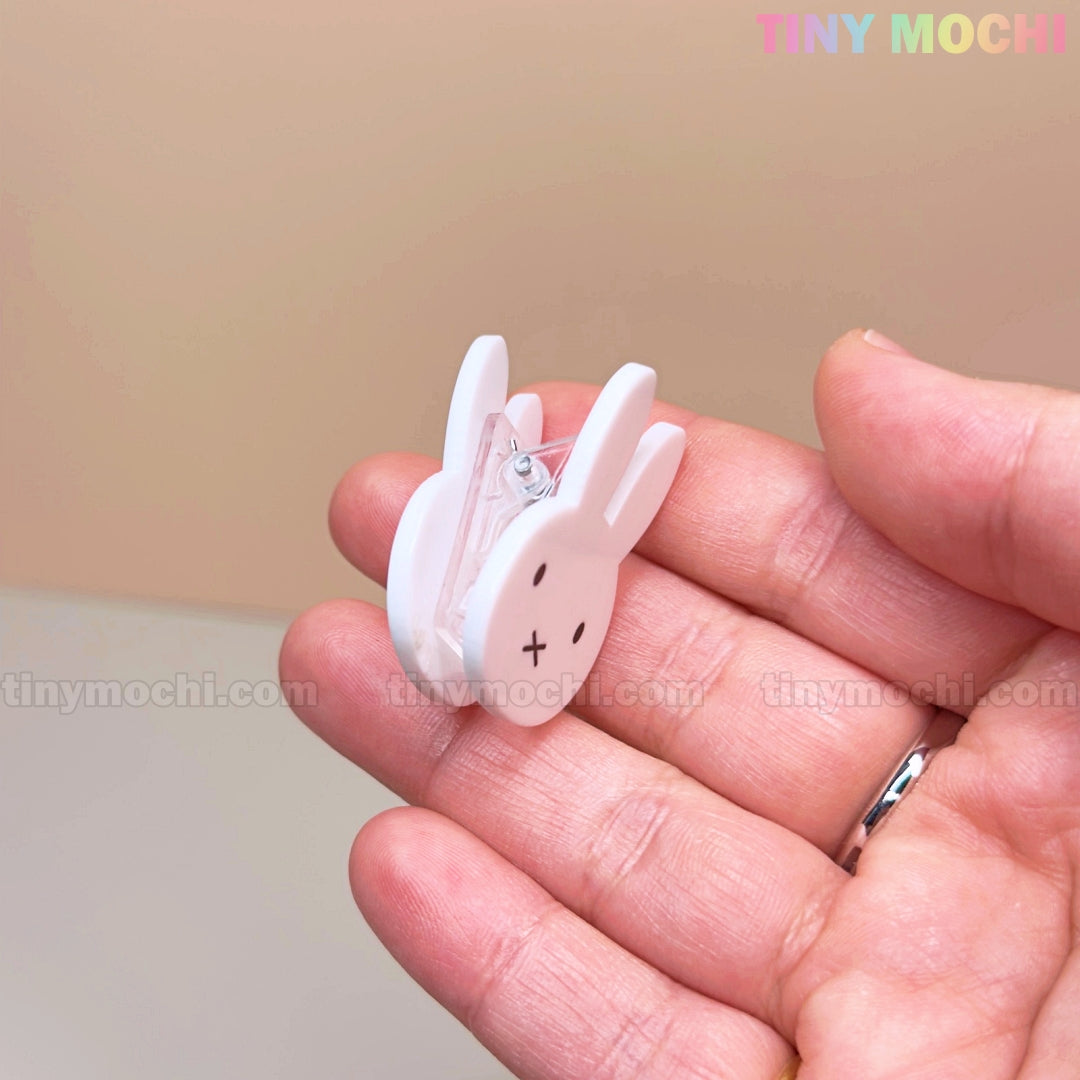 A hand holds the Tiny Mochi Kawaii Bunny Paper Clip—a cute, Miffy-inspired binder clip with black eyes and an X mouth—perfect for desk decor and organization. Set on a soft beige background with “TINY MOCHI” in pastel at the top right.