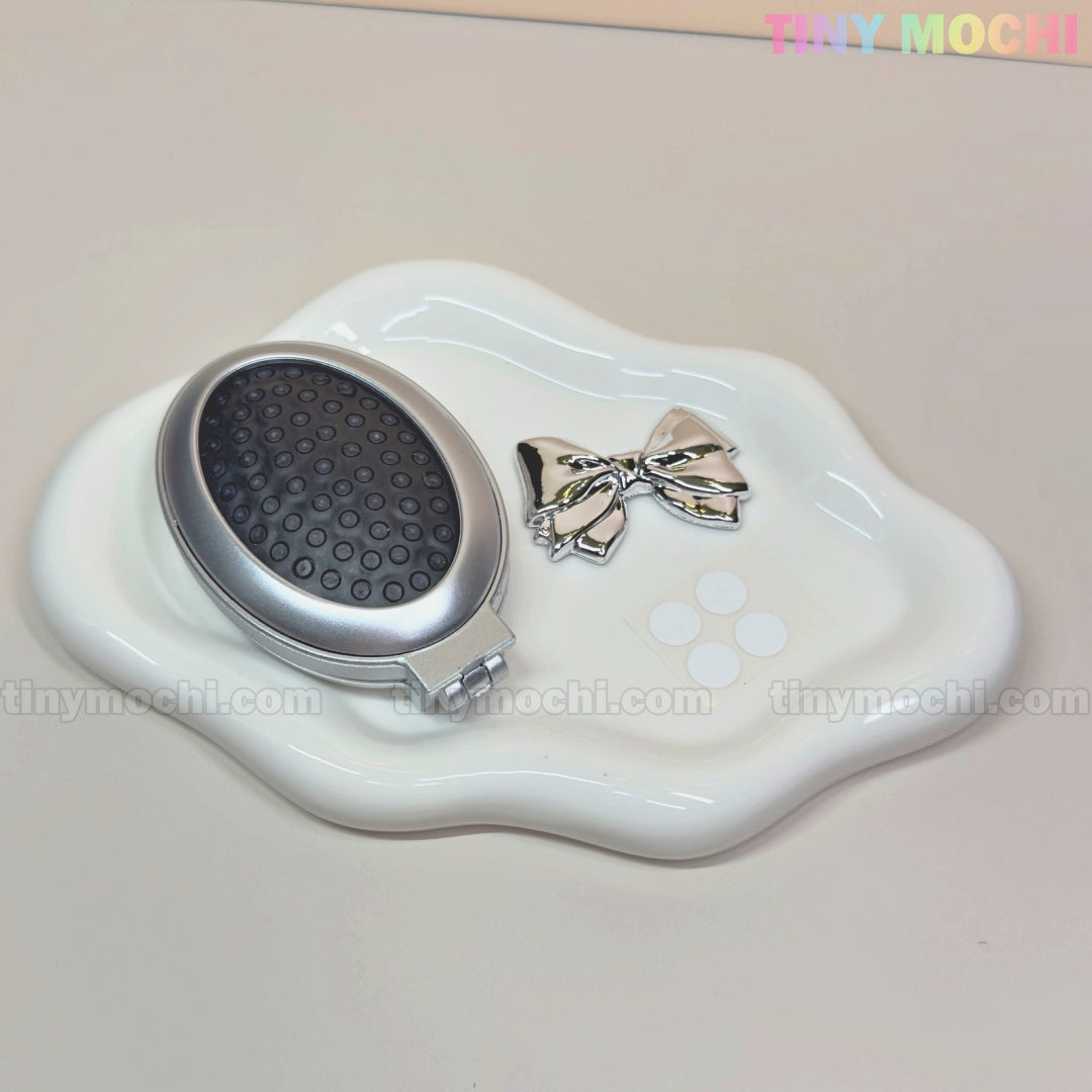 The Tiny Mochi Oval Silver Folding Travel Mirror Comb—a compact pocket-sized brush with built-in mirror—rests on a wavy-edged white dish atop a beige surface, accompanied by a shiny silver bow hair clip.