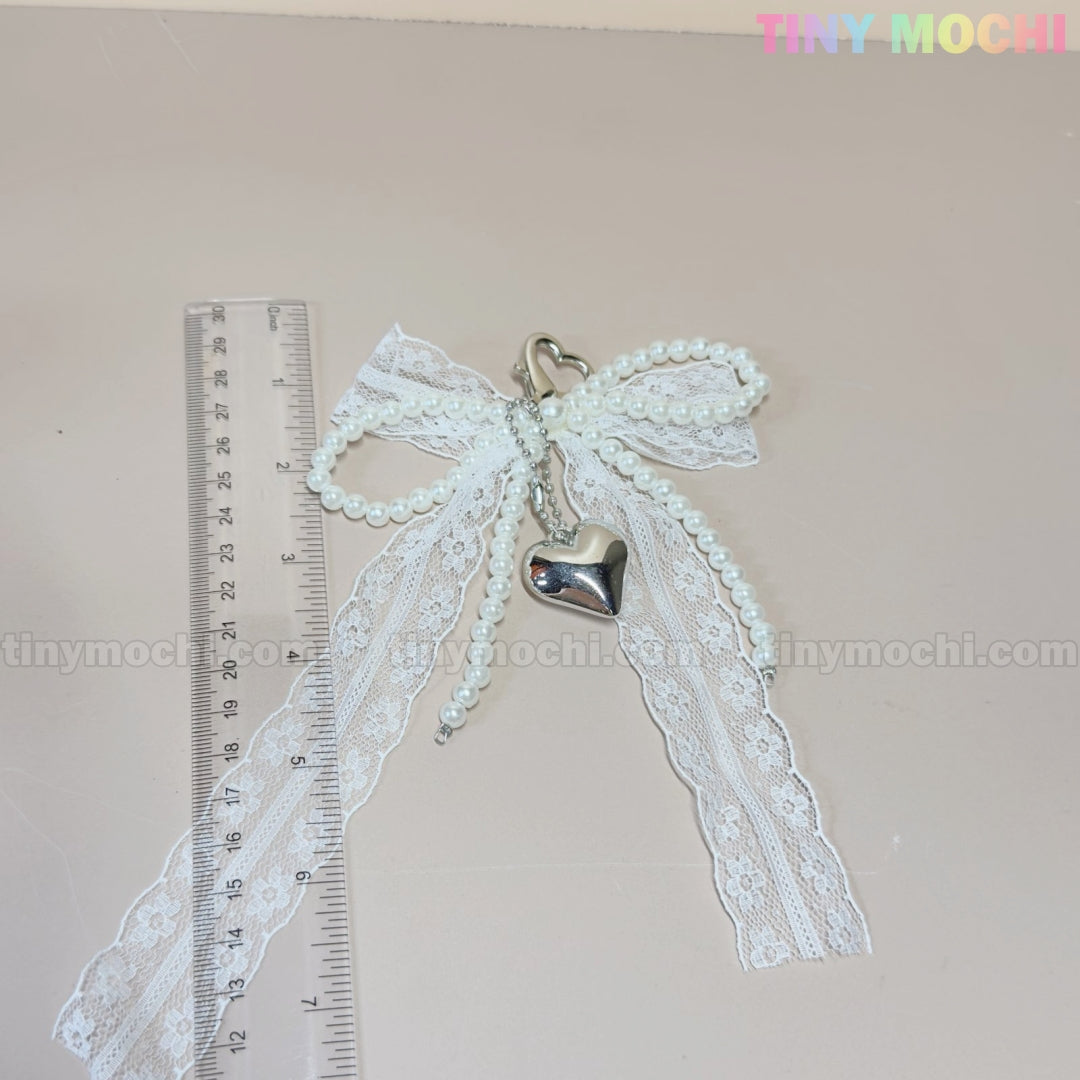 The Pearl Lace Bow Keychain with Silver Heart by Tiny Mochi features faux pearl strands, a heart clasp, and a silver heart charm. Shown on a beige background next to a ruler, “Tiny Mochi” in pastel colors appears at the top right.