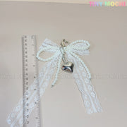 The Pearl Lace Bow Keychain with Silver Heart by Tiny Mochi features faux pearl strands, a heart clasp, and a silver heart charm. Shown on a beige background next to a ruler, “Tiny Mochi” in pastel colors appears at the top right.