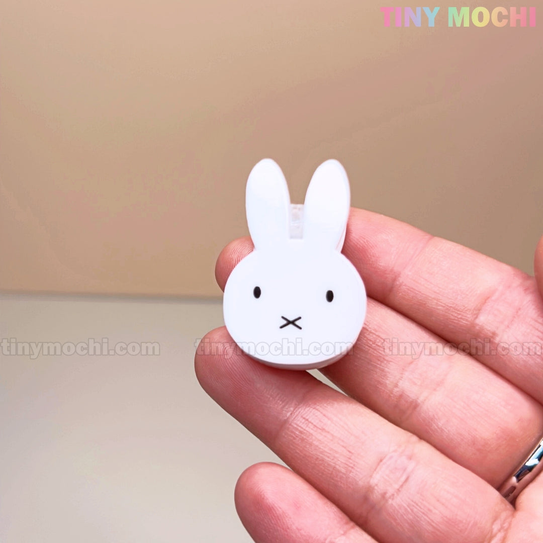 A hand holds the Kawaii Bunny Paper Clip by Tiny Mochi, a Miffy-inspired binder clip with cute black eyes and mouth. This adorable desk accessory features a strong spring clamp. Beige background with TINY MOCHI in pastel at the top right.