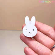 A hand holds the Kawaii Bunny Paper Clip by Tiny Mochi, a Miffy-inspired binder clip with cute black eyes and mouth. This adorable desk accessory features a strong spring clamp. Beige background with TINY MOCHI in pastel at the top right.