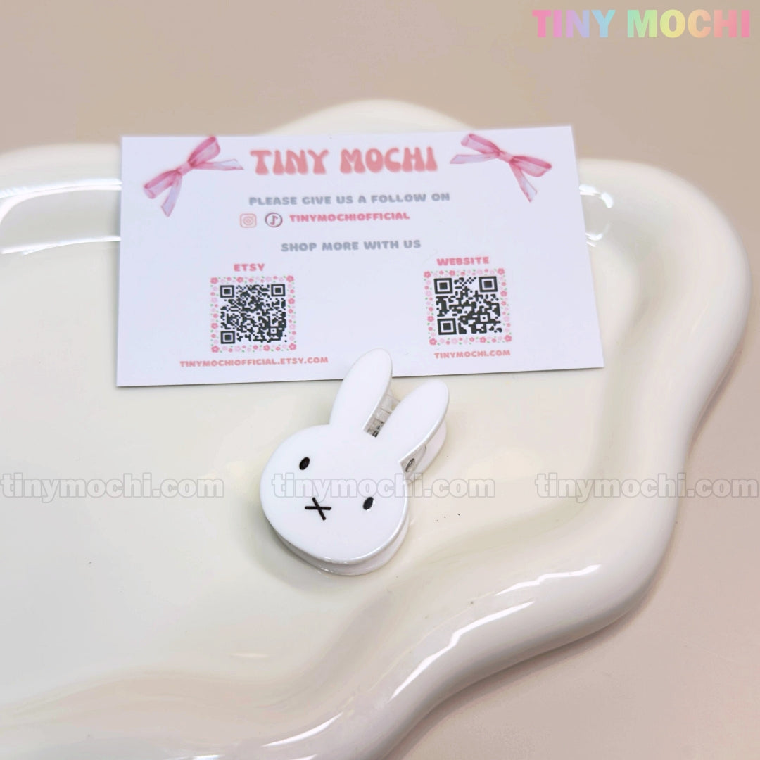 A Kawaii Bunny Paper Clip—Cute Miffy-Inspired Binder Clip by Tiny Mochi—sits on a glossy, wavy-edged ceramic dish with a business card featuring two QR codes and pastel accents, perfect for cute desk decor and organization.