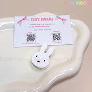 A Kawaii Bunny Paper Clip—Cute Miffy-Inspired Binder Clip by Tiny Mochi—sits on a glossy, wavy-edged ceramic dish with a business card featuring two QR codes and pastel accents, perfect for cute desk decor and organization.