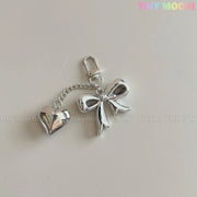 The Silver Bow Heart Keychain – Kawaii Mirror Bag Charm for Backpacks by Tiny Mochi features a shiny heart linked to a large bow clasp, displayed on a light surface. TINY MOCHI appears in pastel at the top right—ideal for any kawaii keychain collection.