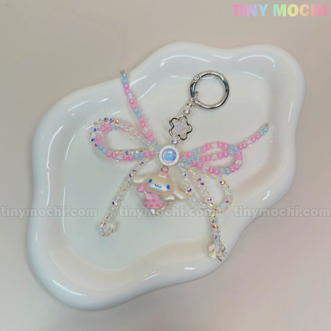 The Cotton Candy Wings Charm Keychain by Tiny Mochi features pink, blue, and clear pastel beads, a bow, and a small bear charm in kawaii fairycore style. Finished with a metallic clasp, it’s a cute phone or bag accessory.